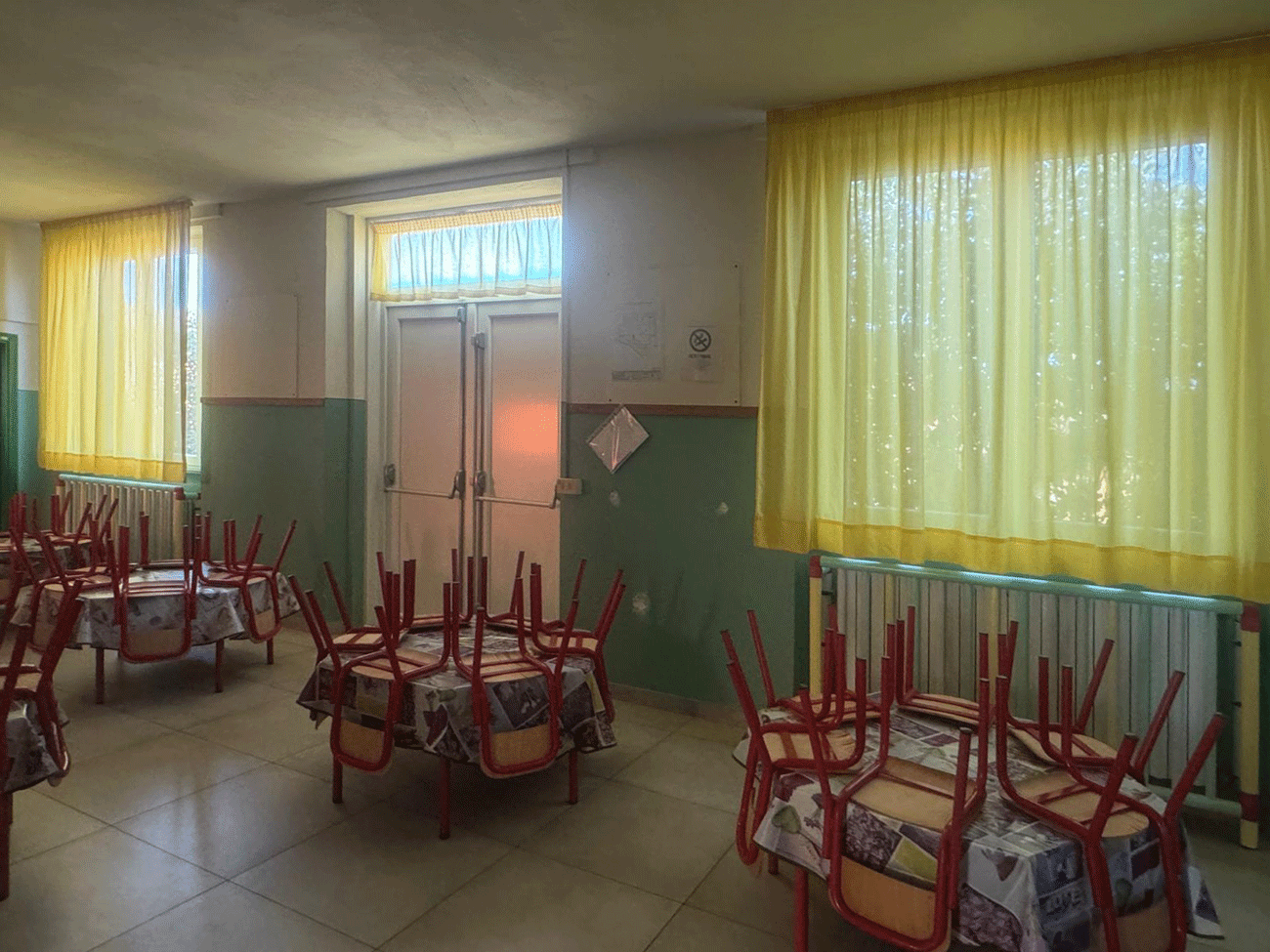 Interno di una mensa o aula con tavolini rotondi, sedie capovolte e tende