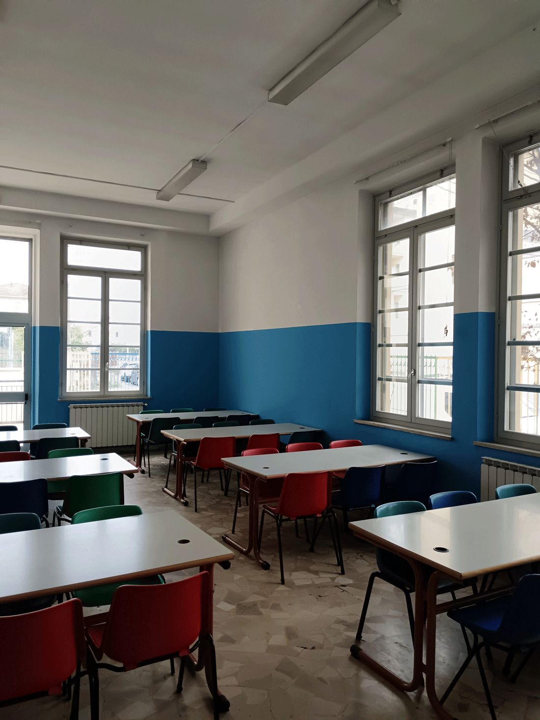 Aula scolastica luminosa con banchi disposti a gruppi e sedie colorate.