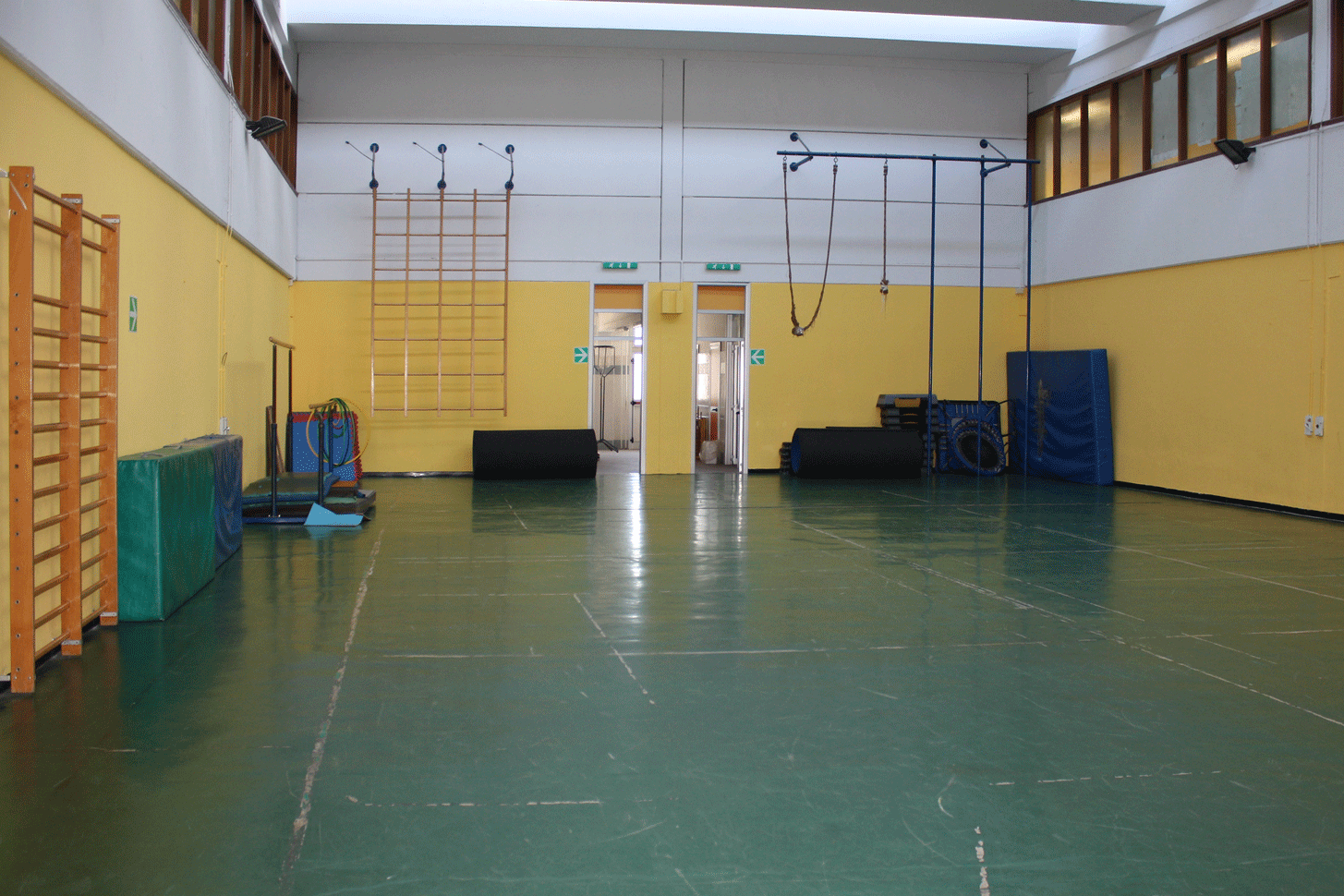 Palestra scolastica con pareti gialle, pavimentazione verde e attrezzature sportive come spalliere e tappeti.