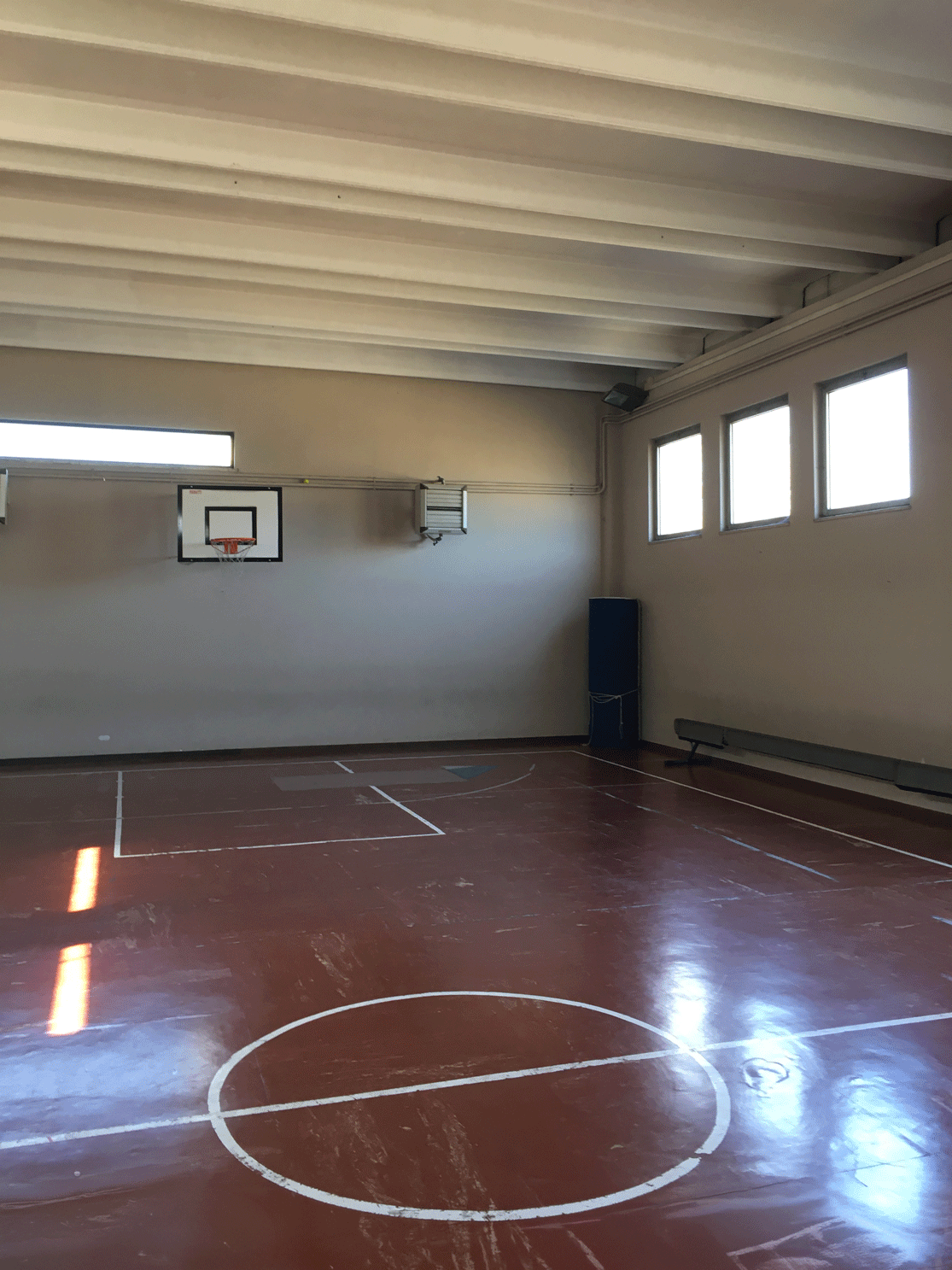 Interno di una palestra con campo da basket, pareti grigie e soffitto con travi a vista.