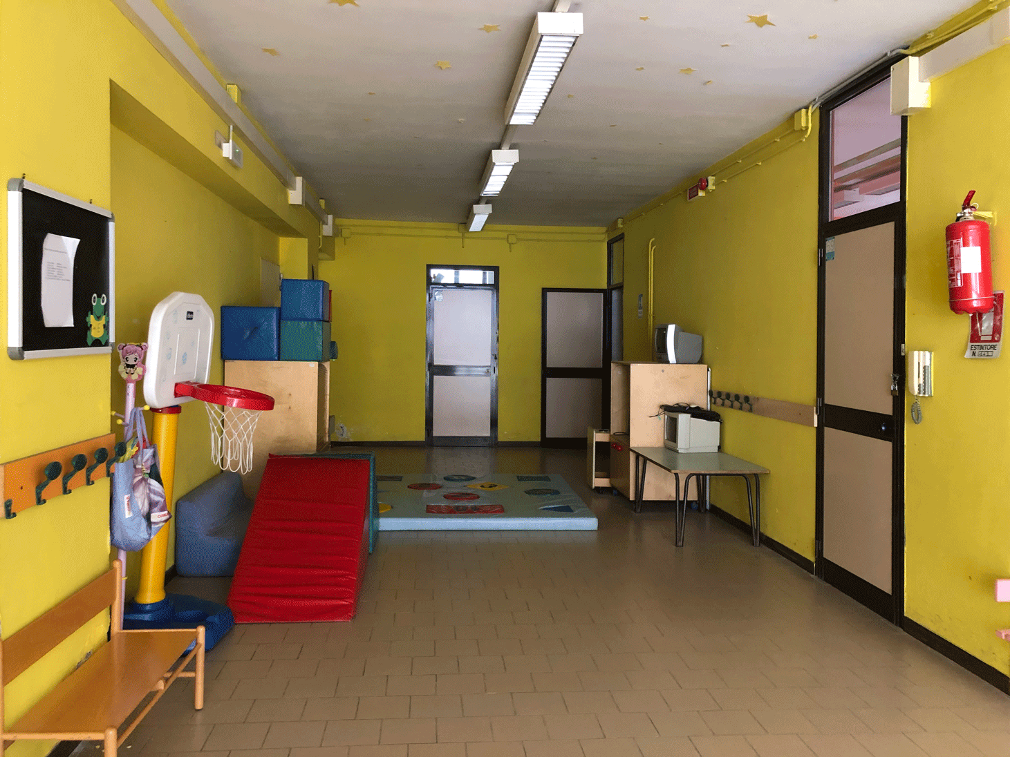 Corridoio scolastico con pareti giallo acceso, giochi per l'infanzia ed estintore a muro ben visibile.