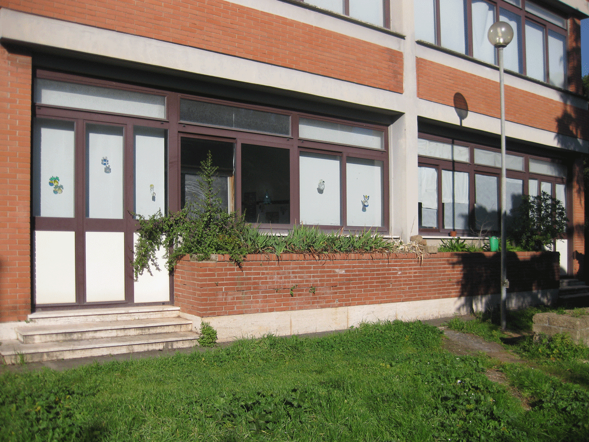 Facciata esterna di un edificio scolastico in mattoni con ampie vetrate e un'aiuola rialzata.