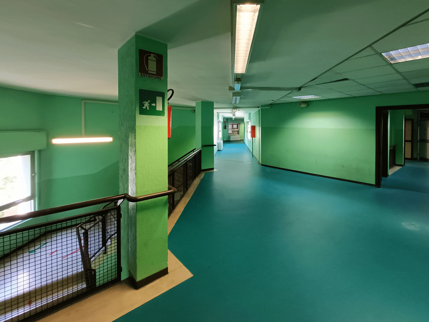 Corridoio scolastico interno con segnaletica antincendio e pavimentazione azzurra.