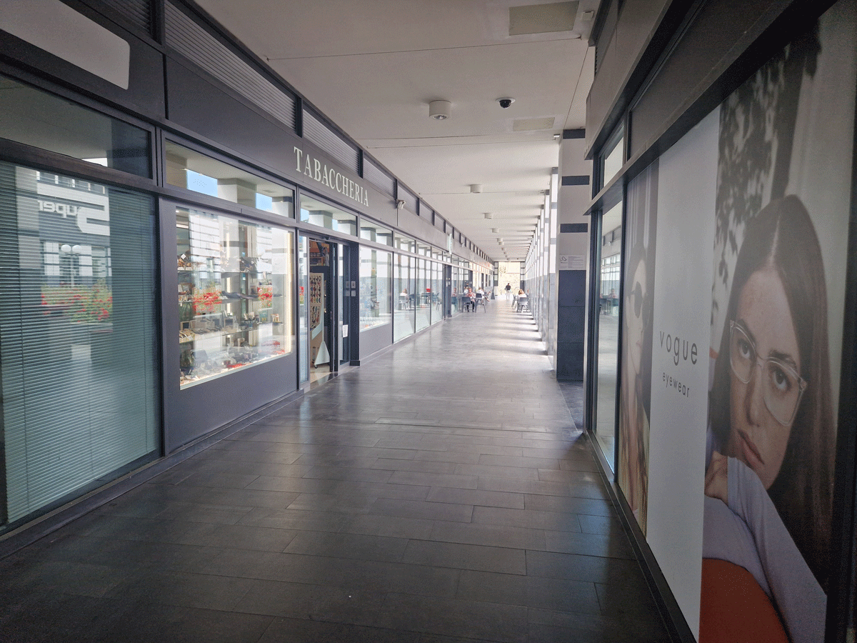 Corridoio della galleria commerciale Esselunga con negozi, tabaccheria e sistemi di illuminazione a soffitto.