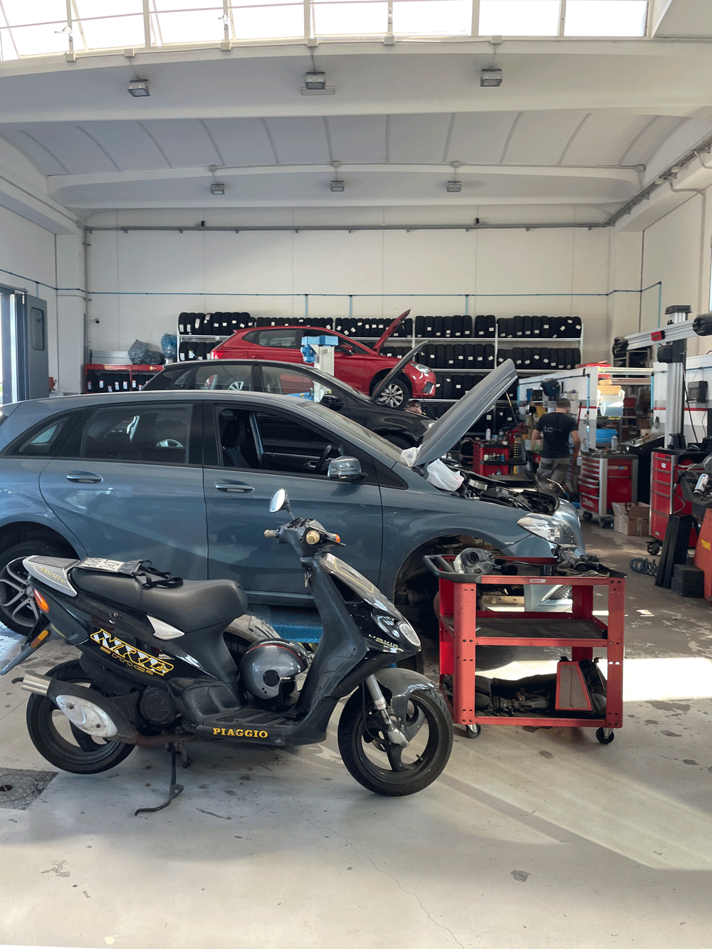 Interno dell'autofficina LC-CAR con auto sui ponti sollevatori, moto e scaffalature di pneumatici sullo sfondo.