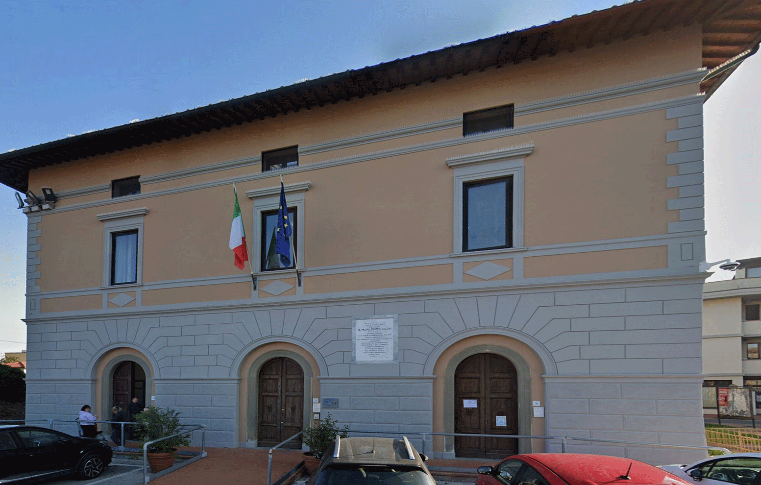 Primo piano della facciata storica degli uffici comunali con bandiere istituzionali e ingressi ad arco.